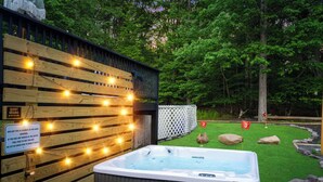 A hot tub next to a wooden deck with string lights, surrounded by a fenced yard with a golf course-like setting. - Heated Pool, Mini Golf, Bowling, Hot Tub, GameRoom (East Stroudsburg)