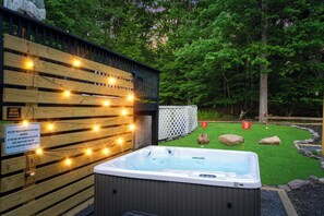 A hot tub next to a wooden deck with string lights, surrounded by a fenced yard with a golf course-like setting. - Heated Pool, Mini Golf, Bowling, Hot Tub, GameRoom (East Stroudsburg)
