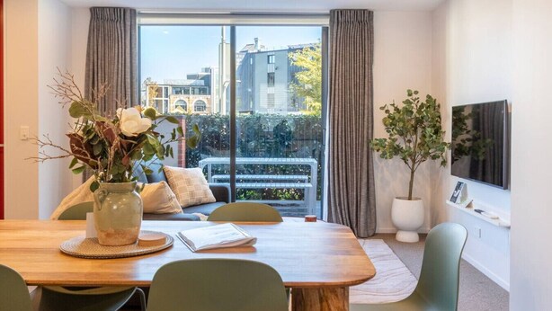 Et moderne værelse med spisebord, stole, sofa, fjernsyn og store vinduer med gardiner.