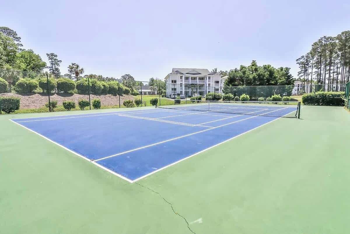Un court de tennis avec un filet, entouré de verdure et avec un bâtiment en arrière-plan.