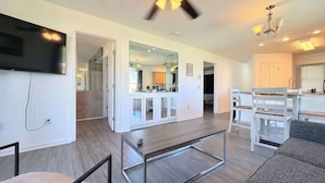 A modern living room with a flat-screen TV, a dining area with white furniture, and a kitchen visible through an open doorway. - Spacious 2BR Getaway in Myrtle Beach, Tennis, Pool (Myrtle Beach)