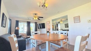 A modern living room with a round wooden table, white chairs, a ceiling fan, and a television mounted on the wall. - Spacious 2BR Getaway in Myrtle Beach, Tennis, Pool (Myrtle Beach)