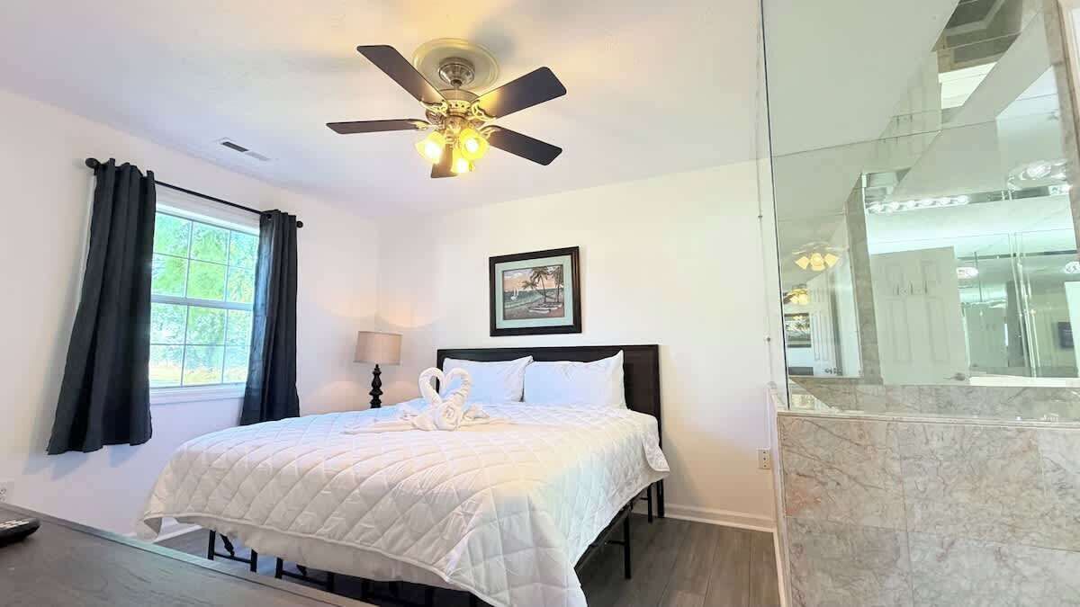 A bedroom with a bed, a ceiling fan, a window with curtains, a framed picture on the wall, and a bathroom with a marble countertop.