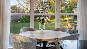 A round table with four chairs, a vase with flowers, and a view of a backyard through large windows. - Forrest Hill Oasis with Pool, Spa and BBQ (Auckland North Shore)