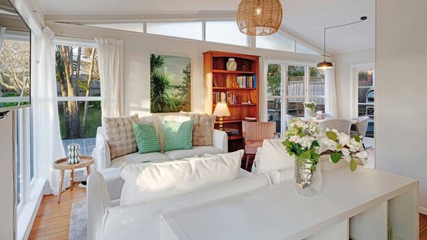 A spacious living room with a white sofa, a bookshelf, and a dining area with a table and chairs. - Forrest Hill Oasis with Pool, Spa and BBQ (Auckland North Shore)