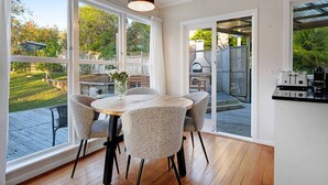 A dining area with a round table and four chairs, large windows, and a view of a backyard with a grill. - Forrest Hill Oasis with Pool, Spa and BBQ (Auckland North Shore)