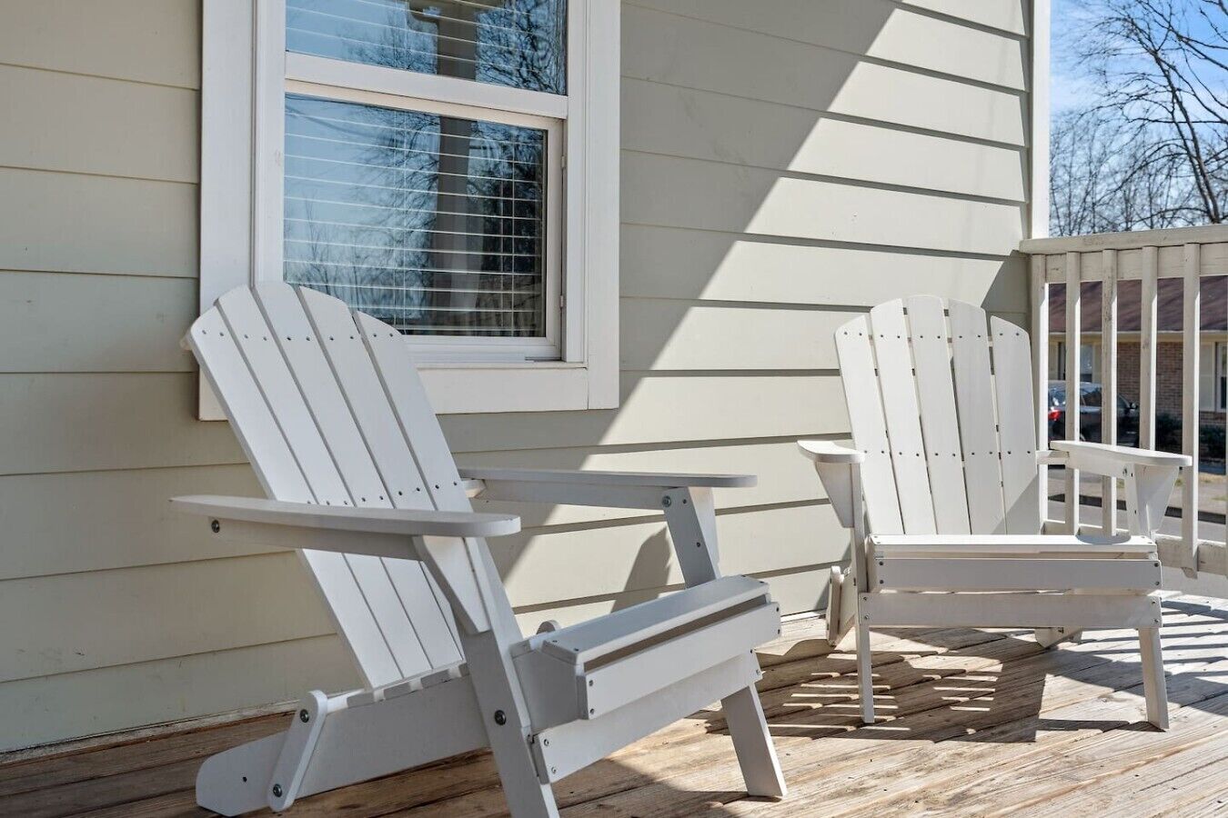 Two white Adirondack chairs on a wooden deck, a window with blinds, and a building with horizontal siding.