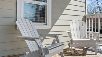 Two white Adirondack chairs on a wooden deck, a window with blinds, and a building with horizontal siding.