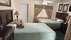 Ein Hotelzimmer mit zwei Betten, einem Schreibtisch, einer Kommode, einem Deckenventilator und einem Fenster mit Vorhängen.