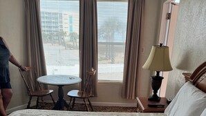 A hotel room with a bed, a round table, two chairs, a lamp, and a view of the outside. - Room 10-Traditional Motel Room w/Oceanview (Daytona Beach)