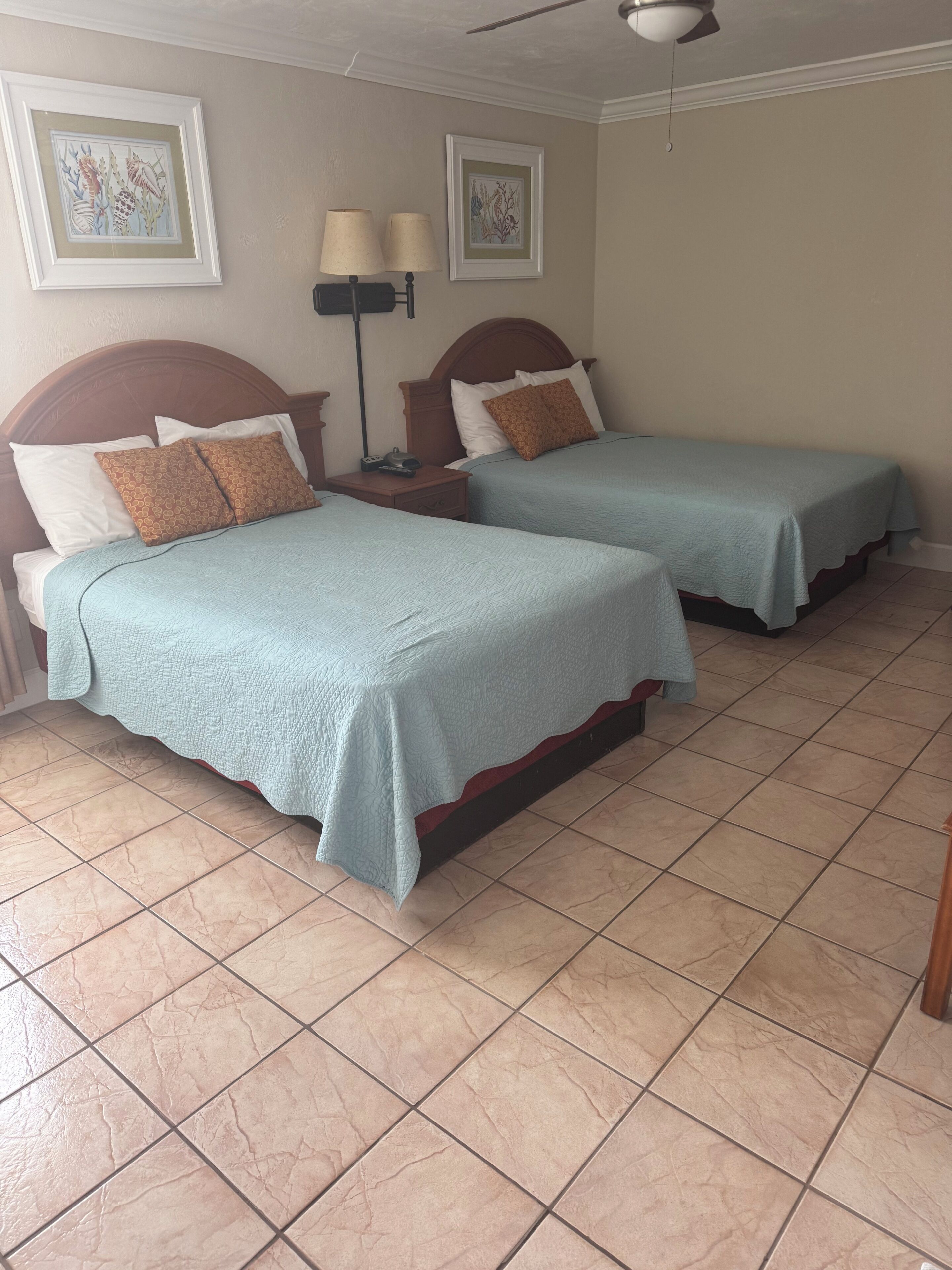 A hotel room with two beds, a nightstand, a lamp, and artwork on the walls.