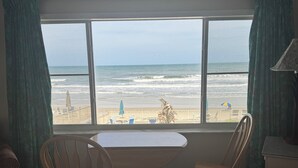 Ein Zimmer mit großem Fenster und Blick auf den Strand, einem Tisch, Stühlen und einer gepolsterten Couch mit Muster.