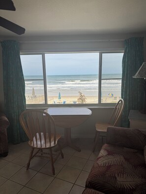 Ein Zimmer mit großem Fenster und Blick auf den Strand, einem Tisch, Stühlen und einer gepolsterten Couch mit Muster.
