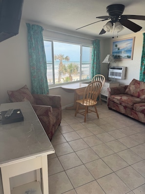A room with a dining table, a sofa, a chair, a ceiling fan, a window with a view of the ocean, and an air conditioning unit. - Room 19-Efficiency Unit-Ocean Front 1st floor (Daytona Beach)