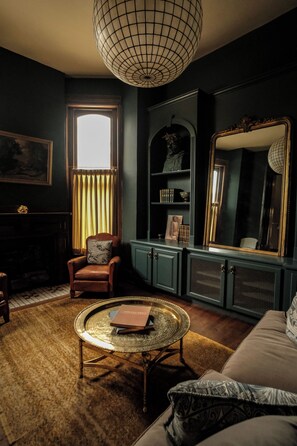 Ein Zimmer mit einem runden Couchtisch, einer Couch, einem Sessel, einem Kamin, einem Bücherregal und einem großen Spiegel.