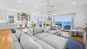 A spacious living area with a large gray sectional sofa, a dining table with chairs, and a view of the ocean through large windows. - Beachfront Bliss with Marsh Views & Private Dock (East Sandwich)