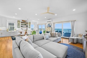 A spacious living area with a large gray sectional sofa, a dining table with chairs, and a view of the ocean through large windows. - Beachfront Bliss with Marsh Views & Private Dock (East Sandwich)