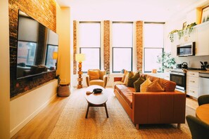 A modern living room with a sofa, a coffee table, a TV mounted on a brick wall, and a kitchen area visible through a large window.