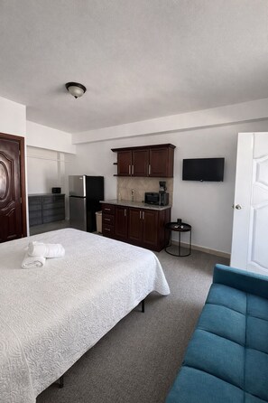 A bedroom with a bed, a blue chair, a kitchenette with cabinets and appliances, and a TV mounted on the wall. - all-inclusive hotel room (cabo rojo)