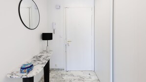 Un couloir avec une porte blanche, un miroir à cadre noir, une console surmontée d’un plateau en marbre et un petit sac aux motifs bleus et rouges.