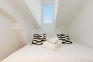 Ein kleiner, weiß gestrichener Raum mit schrägem Dach, einem Bett und zwei Kissen mit Muster, sowie einem Fenster.