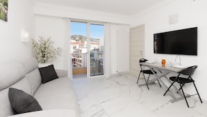 Una sala moderna con un sofá, una mesa de comedor con sillas, un televisor montado en la pared y un ventanal corredero que da a un balcón.