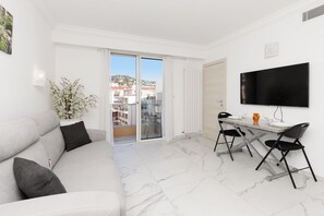 A modern living room with a sofa, a dining table with chairs, a TV mounted on the wall, and a sliding glass door leading to a balcony. - Upstays  Sea View 25m² Studio Moure Rouge (Cannes)