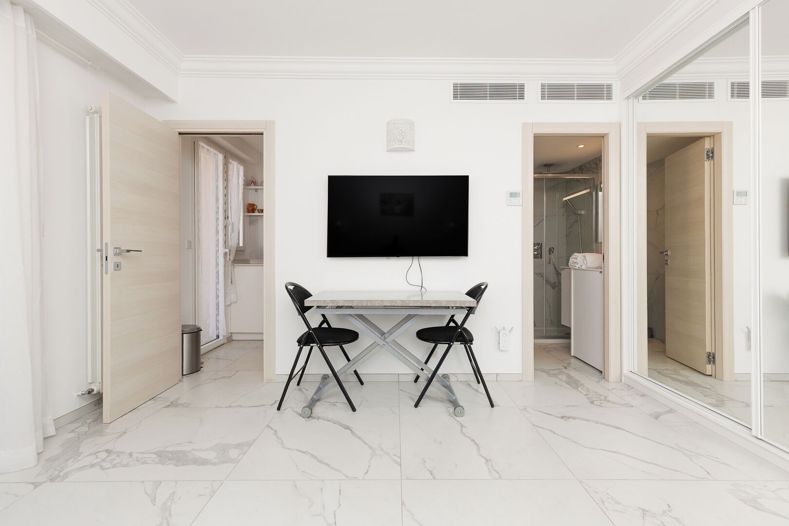 A modern hotel room with a flat-screen TV mounted on the wall, a dining table with chairs, and a marble floor.