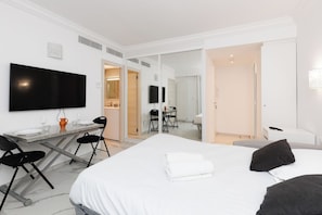 A hotel room with a bed, a desk, a chair, a TV, and a bathroom visible through an open door. - Upstays  Sea View 25m² Studio Moure Rouge (Cannes)