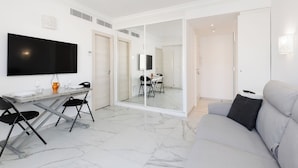 Una sala moderna con un sofá, una mesa de comedor con sillas, un televisor de pantalla plana y un ventanal corredero que da a otra habitación.