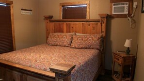A bedroom with a wooden headboard, a patterned bedspread, a nightstand with a lamp, and a wall-mounted air conditioning unit. - Riverfront Cabins  and  Vacation Homes (Rio Frio)