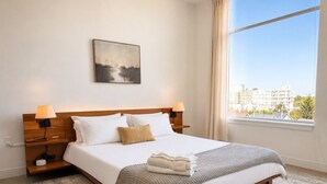 A neatly made bed with a blanket and pillows, a bedside table with a lamp, a wall-mounted painting, and a large window with a city view. - Bay View Designer 2BR · Best Location with Balcony, Gym, and Terrace · Sleeps 6 (Milwaukee)