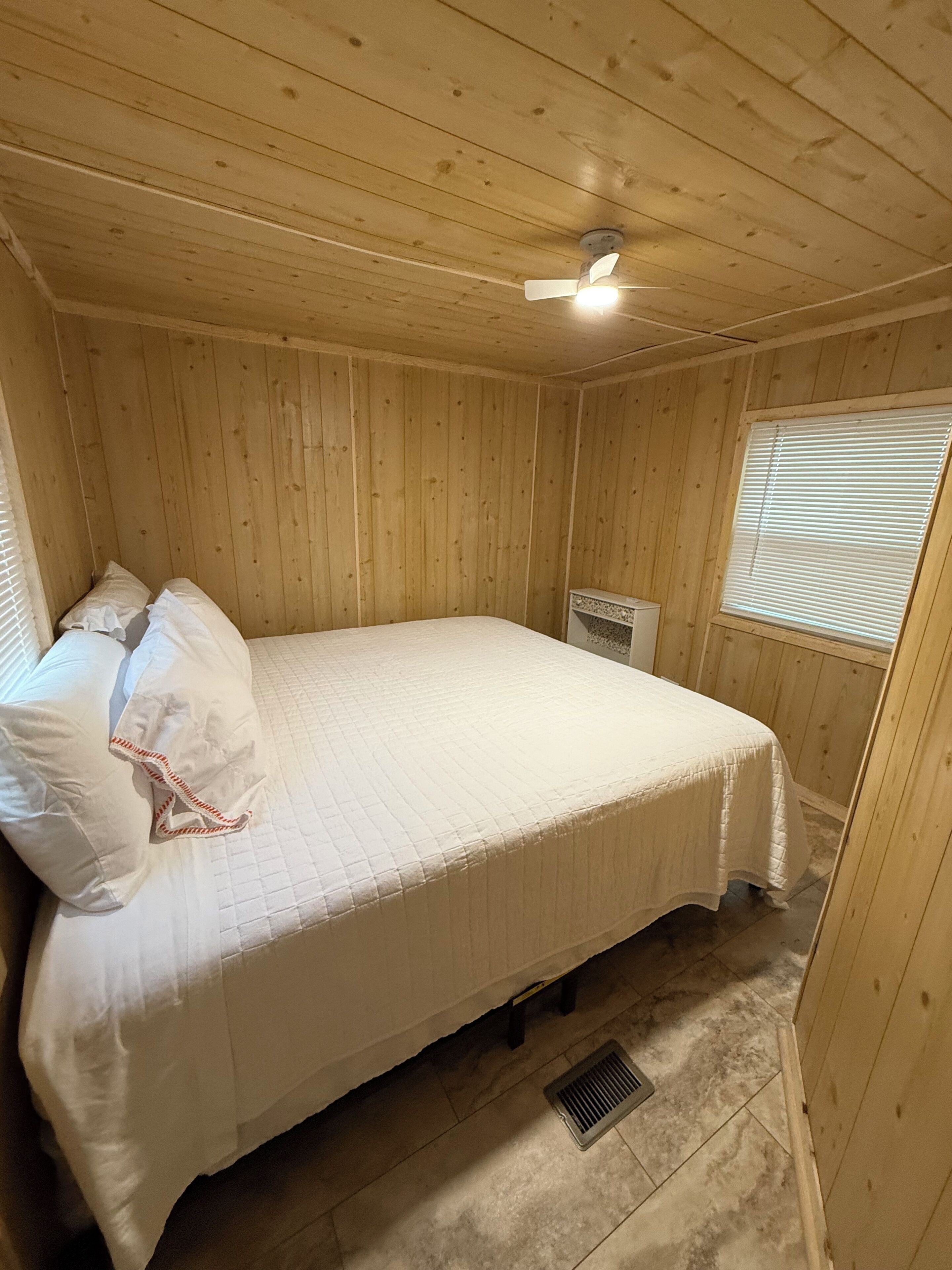 A wooden-paneled room with a bed, a ceiling fan, a window with blinds, and a vent.