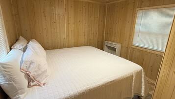 A wooden-paneled room with a bed, a ceiling fan, a window with blinds, and a vent.