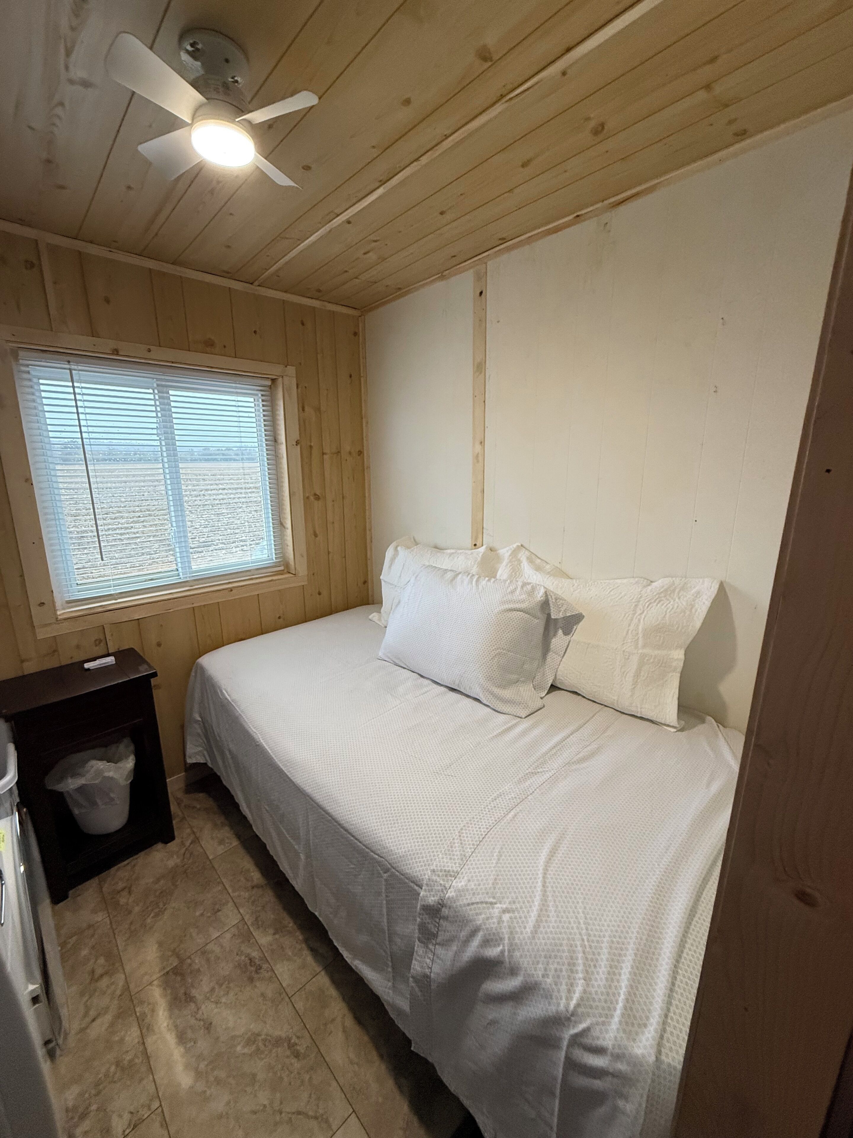A bedroom with a bed, a ceiling fan, a window with blinds, a nightstand, and a trash bin.