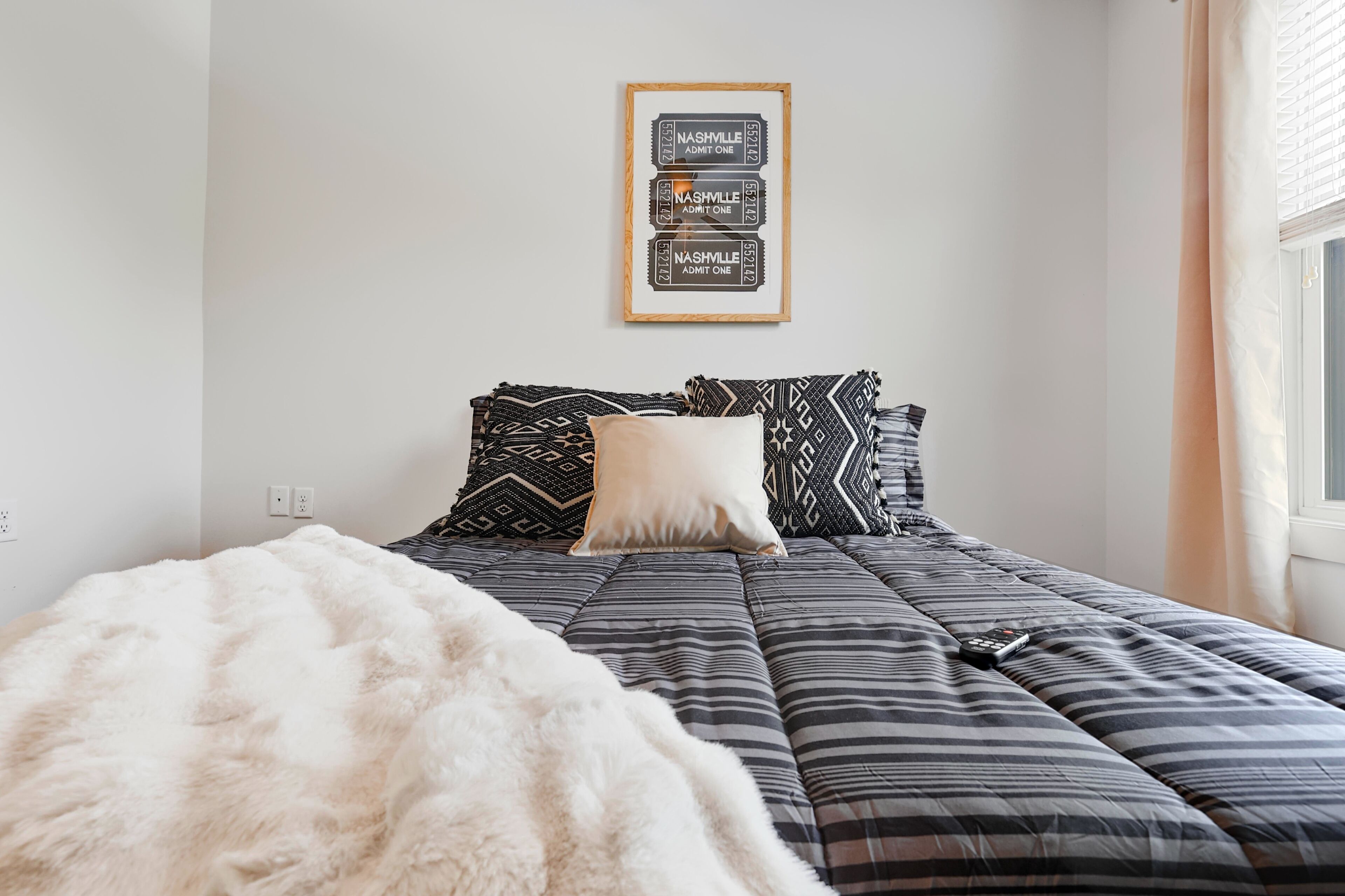 A bedroom with a bed, a fluffy white blanket, patterned pillows, and a framed ticket artwork on the wall.