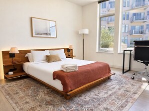 Un cuarto de hotel ordenado con cama, lámparas de noche, un escritorio y un ventanal grande.
