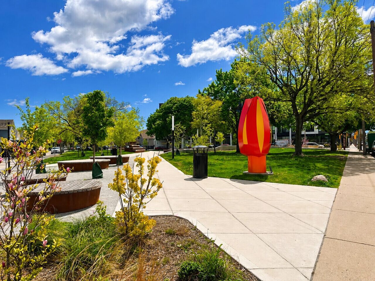 Un parque con una gran escultura roja y amarilla, un camino pavimentado y árboles verdes.