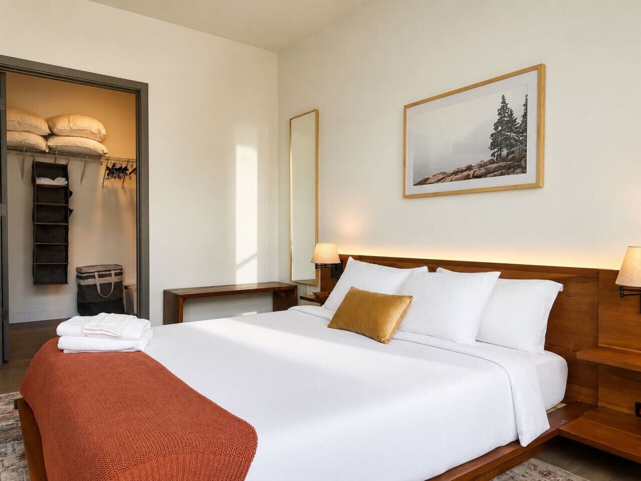 A hotel room with a large bed, a wooden headboard, a nightstand with a lamp, a wall-mounted mirror, and a framed picture of a mountain landscape.