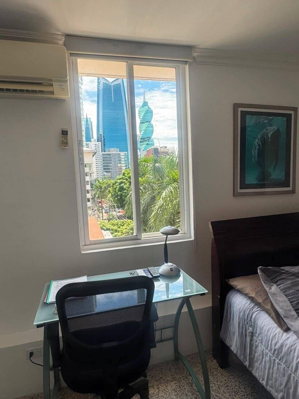 A bedroom with a bed, a desk with a chair, a window with a view of buildings and trees, and a framed picture on the wall.