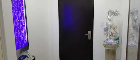 A hallway with a black door, a wall-mounted TV with a blue light, and a decorative silver statue on a pedestal.