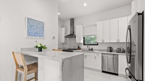 Una cocina moderna con gabinetes blancos, una isla de mármol, electrodomésticos de acero inoxidable y una ventana con persianas.