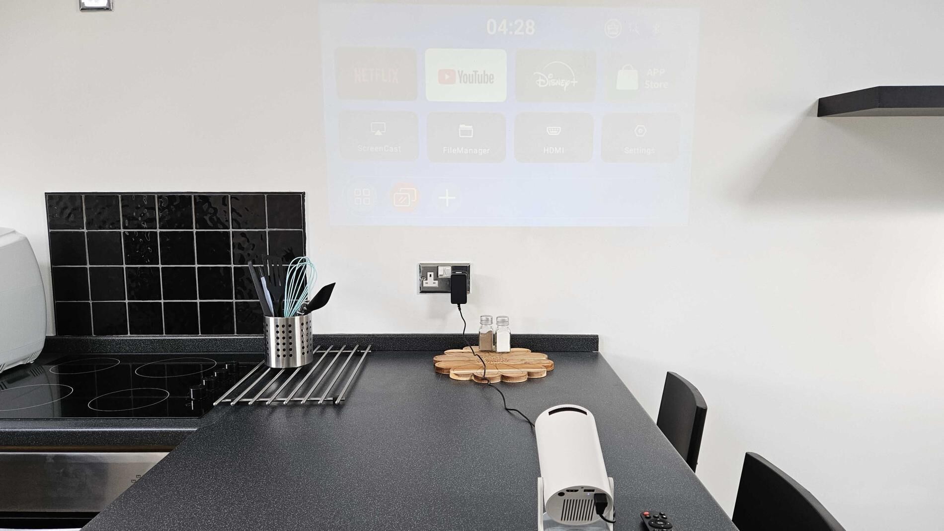 Eine moderne Küche mit schwarzem Arbeitsplatz, einem Projektor, der App-Symbole anzeigt, einer Steckdose und einer Kaffeemaschine.