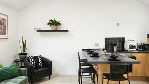 A modern kitchen with a dining area, a black sofa, a green plant, and a small shelf with a plant.