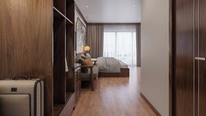 A hotel room with a wooden floor, a bed, a desk with a lamp, a wardrobe, and a suitcase.