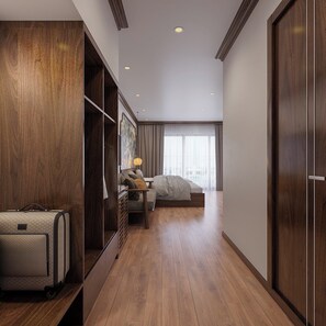 A hotel room with a wooden floor, a bed, a desk with a lamp, a wardrobe, and a suitcase. - Aquarius Garden (Gia Vien)