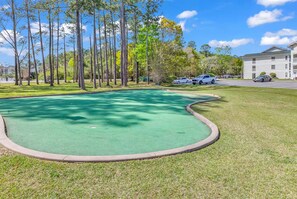 A golf course with a putting green, surrounded by trees and a building in the background. - Golf View River Oaks Condo Pool Myrtle Beach (Myrtle Beach)
