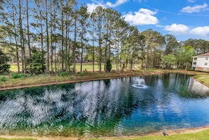 A tranquil pond with a fountain, surrounded by tall trees and a building with a white facade. - Barefoot Landing Condo Pool & Golf View (North Myrtle Beach)