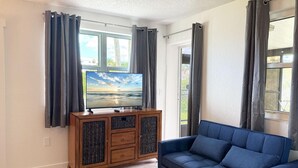A living room with a TV on a wooden cabinet, a blue sofa, and a window with curtains. - Venice Villas Combo – Sleeps 5 - Poolside (Venice)