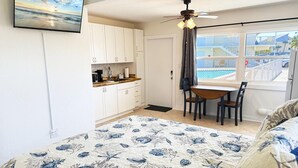 A bedroom with a bed, a ceiling fan, a window with a view of a pool and buildings, and a kitchen area with cabinets and a countertop. - Venice Villas Combo – Sleeps 5 - Poolside (Venice)